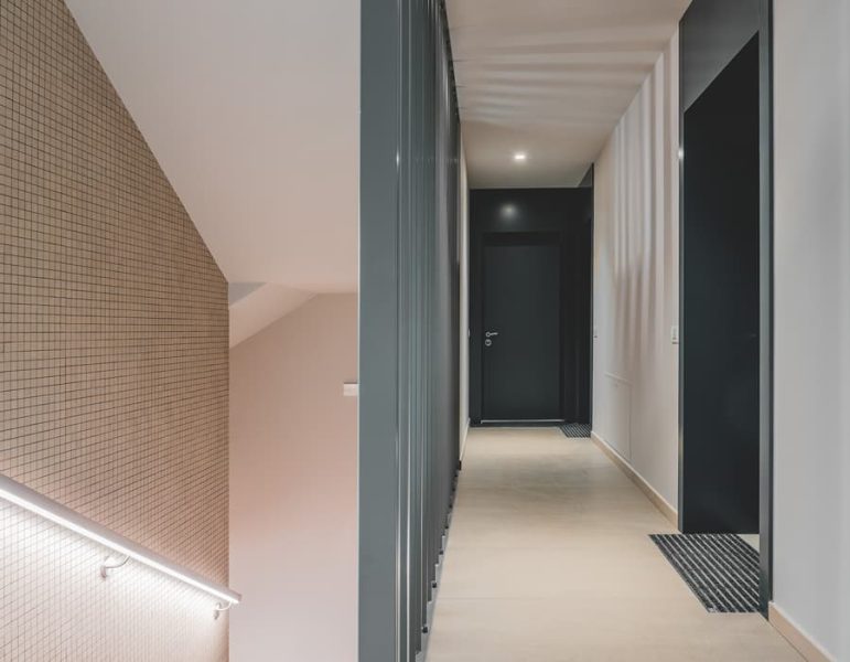 Corridoio interno d'albergo, luci soffuse, pareti chiare e corrimano illuminato, porta nera all’estremità.