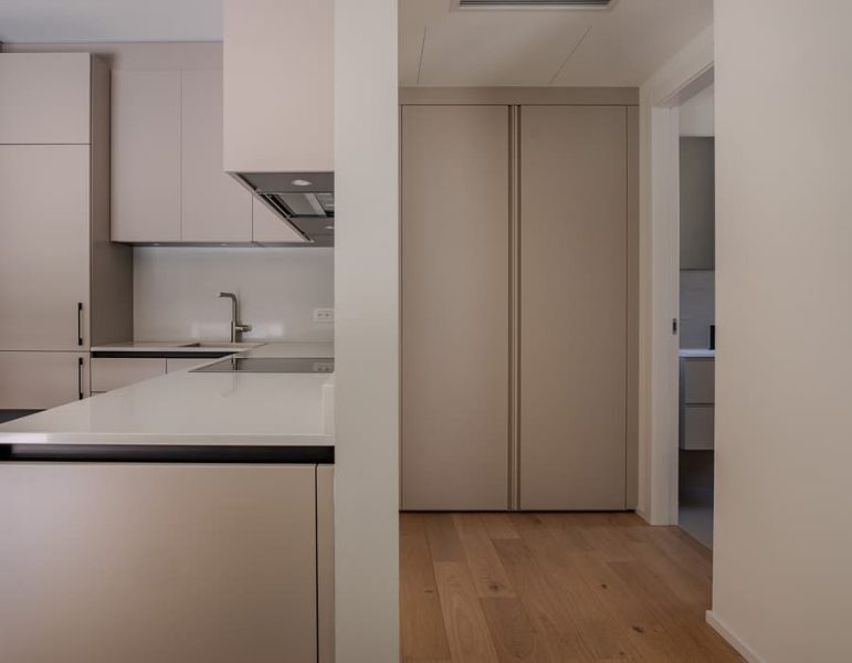 Cucina moderna e minimale con lavello, piano bianco e mobili chiari; spazio aperto e illuminato, con pavimento in legno chiaro e una chiusura scorrevole.