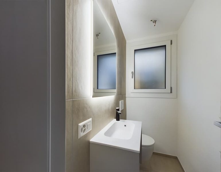 Bagno moderno con lavandino sospeso, specchio con luci a LED, finestra con vetro opaco e rubinetto nero su piano lavabo bianco.