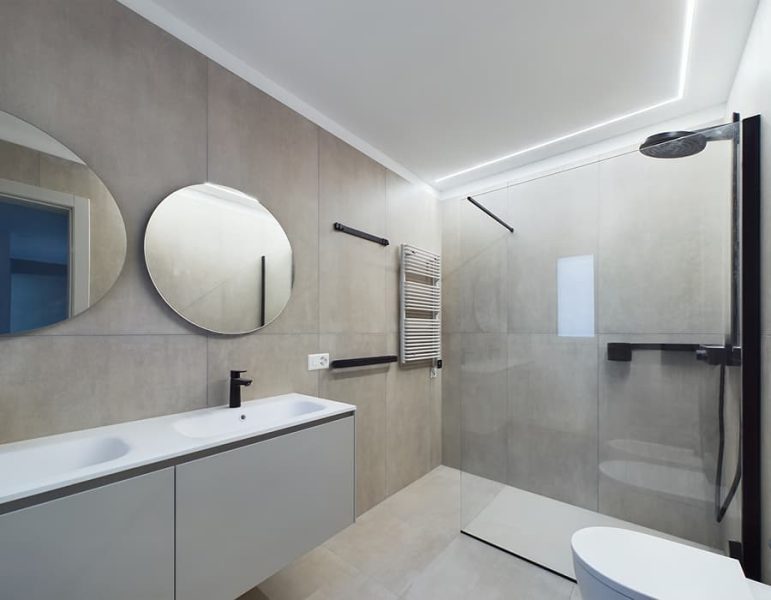 Bagno moderno con doppio lavandino, due specchi rotondi, asciugamano a parete e zona doccia walk-in con soffione fissante nero, tonalità neutre chiare.